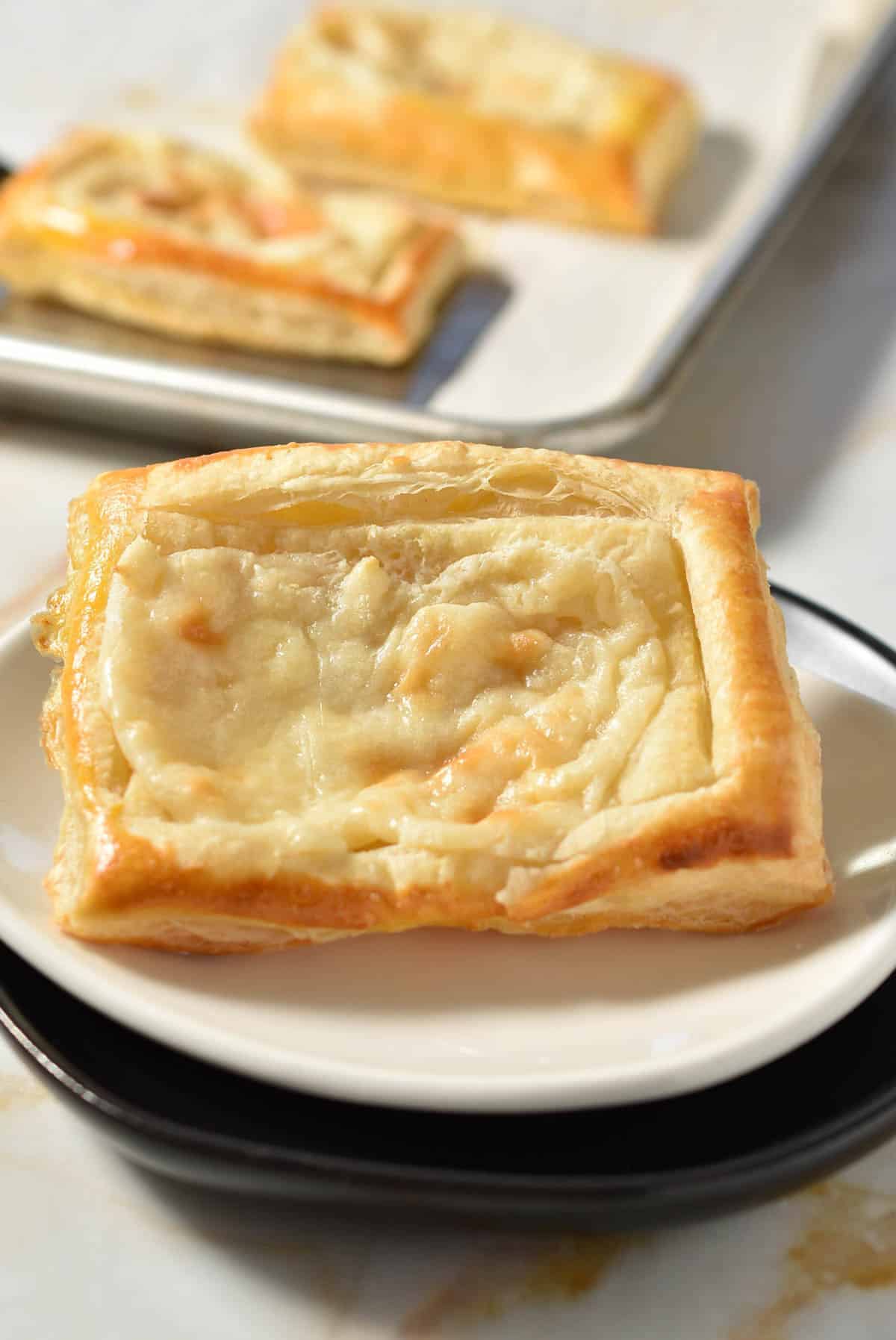A Cheese Danish from Starbucks-a rectangular piece of puff pastry topped with melted cheese-sits on a white plate, with more pastries in the background.