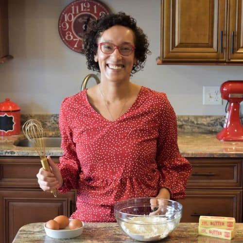 Heather in a red polka dot top stands in a kitchen, holding a whisk beside a bowl of flour, eggs, and butter on the counter.