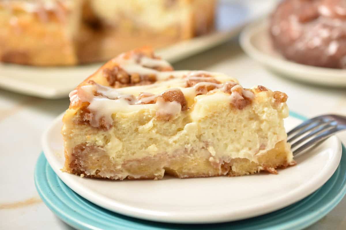 A slice of Honey Bun Cheesecake with cinnamon swirls and icing sits on a white plate, with a fork beside it.