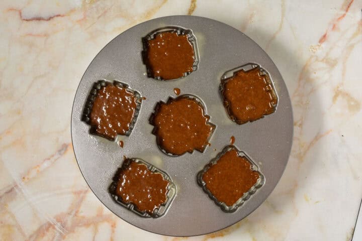 A round bundt pan with six house-shaped molds filled with gingerbread batter, placed on a marbled countertop.