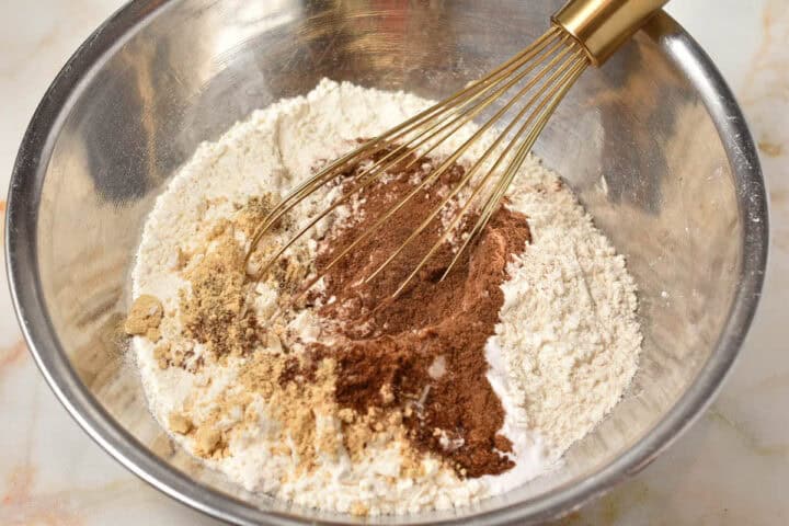 A metal mixing bowl containing flour, cocoa powder, and other dry ingredients for a Gingerbread House Bundt Cake being mixed with a whisk.