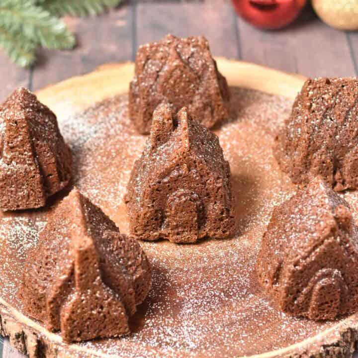 Six Gingerbread House Bundt Cakes, shaped like charming little houses, are arranged on a wooden serving board and dusted with powdered sugar.