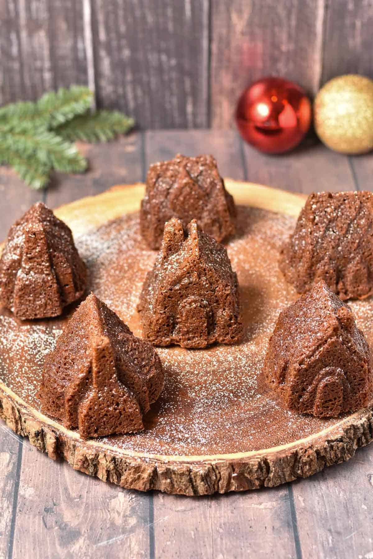 Mini gingerbread cakes shaped like houses and trees are arranged on a wooden platter, dusted with powdered sugar, with holiday ornaments and greenery in the background.