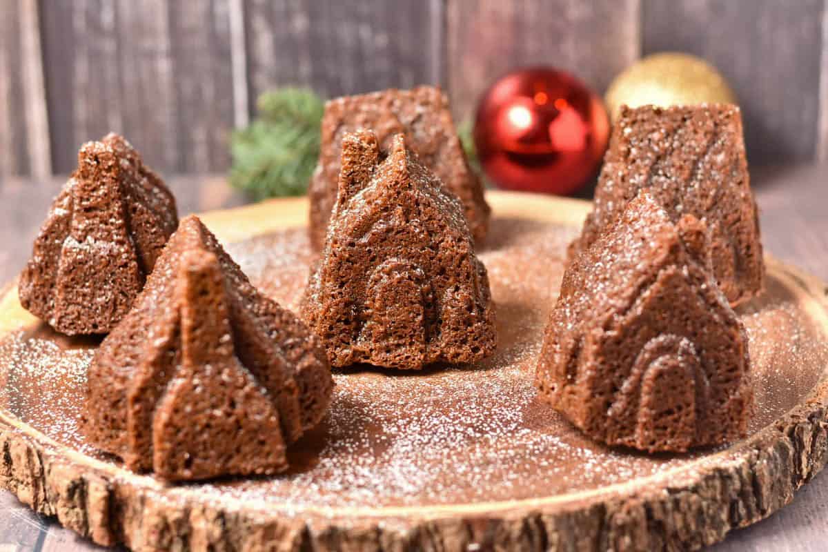 Mini Gingerbread House Bundt Cakes shaped like houses and churches are arranged on a wooden platter, lightly dusted with powdered sugar, with Christmas ornaments in the background.