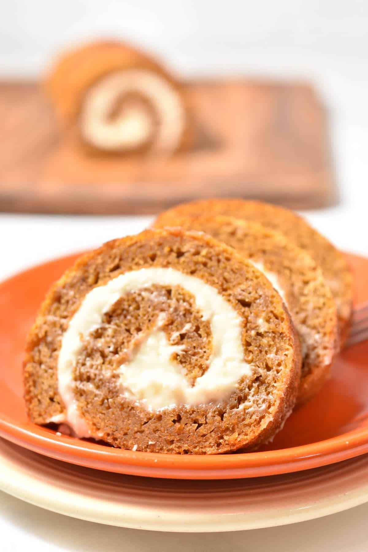 Two slices of pumpkin roll with cream filling are served on an orange plate, with the rest of the roll on a wooden board in the background.