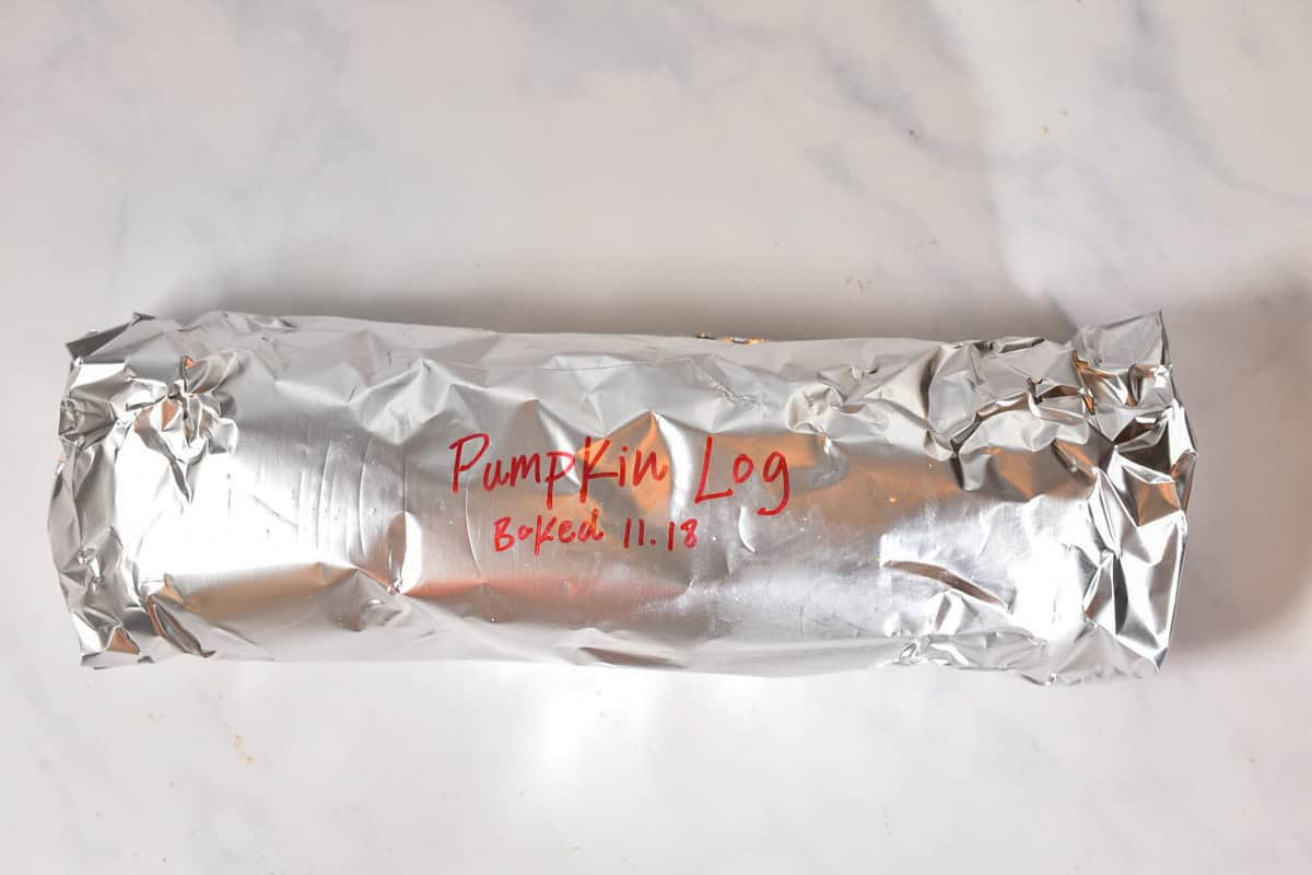 A foil-wrapped roll labeled “Pumpkin Log, Baked 11.18” in red marker sits on a white marble surface—perfect as a Make-Ahead Pumpkin Roll for your Thanksgiving dessert table.