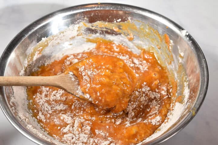 A metal bowl with partially mixed orange batter-perfect for a Make-Ahead Pumpkin Roll-flour visible, and a wooden spoon stirring the mixture, ready for your Thanksgiving baking.
