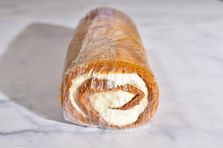 A Pumpkin Roll cake with cream cheese filling is wrapped in clear plastic wrap and placed on a white marble surface-perfect as a make-ahead Thanksgiving dessert.