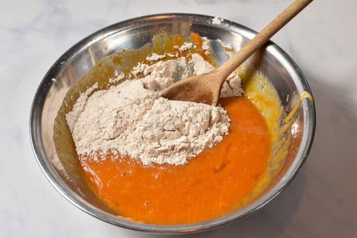 A metal mixing bowl with orange Pumpkin Roll batter and a mound of flour, being stirred with a wooden spoon-perfect for Thanksgiving or as a make-ahead dessert.