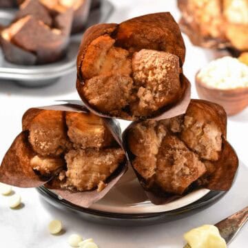 Three cinnamon crumb muffins in brown paper wrappers are stacked on a white plate, with more muffins and baking ingredients in the background-perfect for fans of Starbucks Copycat Cinnamon Pull-Apart Bread.