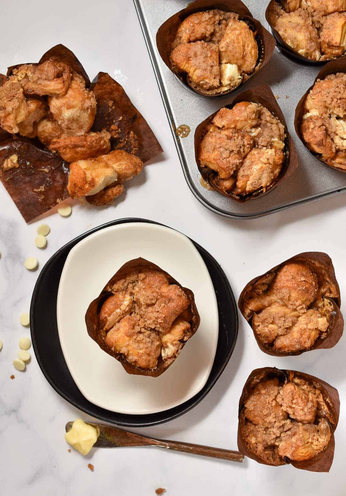 Overhead view of several cinnamon streusel muffins in brown wrappers, some on a plate, others in a muffin tinโreminiscent of Starbucks Copycat Cinnamon Pull-Apart Breadโwith white chocolate chips and a buttered knife nearby.