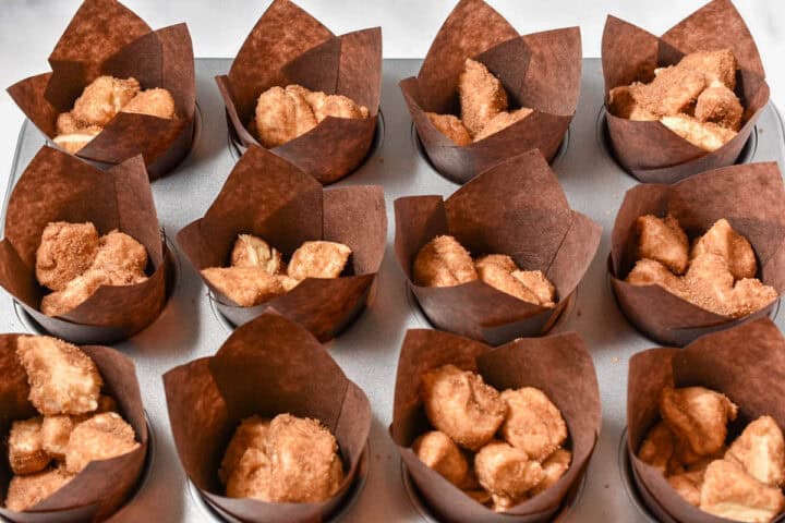 A muffin tin filled with twelve brown paper liners, each containing pieces of cinnamon sugar-coated dough, perfect for making Starbucks Copycat Cinnamon Pull-Apart Bread at home.